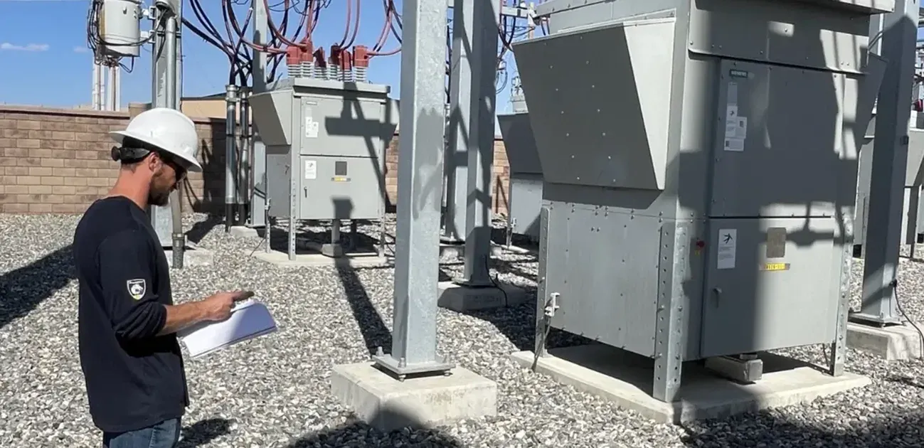 A person wearing a white hard hat and safety glasses holds a clipboard while inspecting equipment at an outdoor electrical substation surrounded by gravel and metal structures.