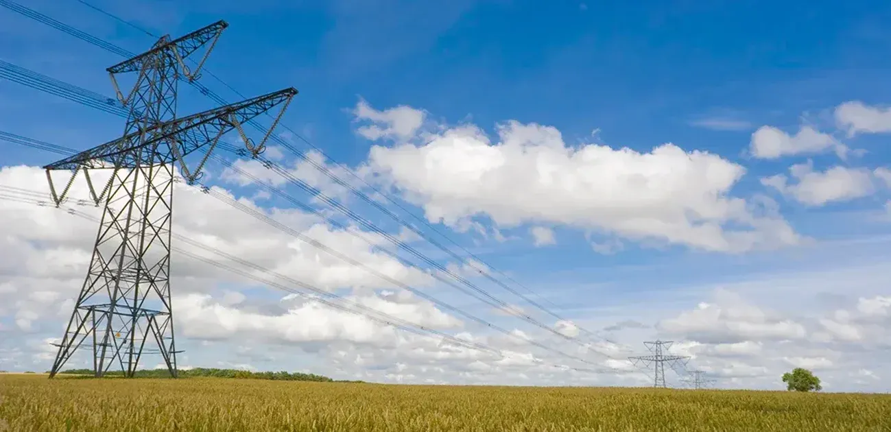 High-voltage power lines run across a wheat field under a blue sky with scattered clouds. The metal towers stretch into the distance, and the golden wheat covers the foreground.