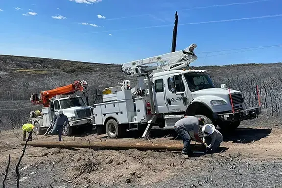 Utility workers install a new wooden pole next to two service trucks in a barren, recently burned landscape under a clear sky.